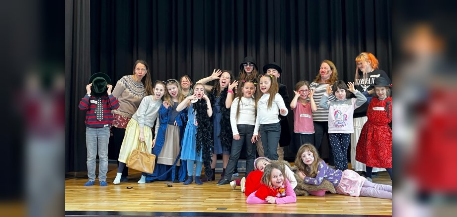 Das Foto zeigt die Teilnehmer des Schau-Spiel-Tages. Sie stehen geschminkt und kostümiert nebeneinander auf der Bühne der Göttenbach-Aula und schneiden Grimassen.