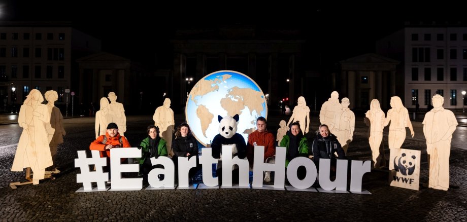 Im Jahr 2025 nahmen in Deutschland über 500 Städte an der Earth Hour des WWF teil. (Foto: ) Das Foto zeigt mehrere Personen und Pappmännchen auf einem dunklen Platz. Sie sitzen hinter einem Schriftzug "Earth Hour".