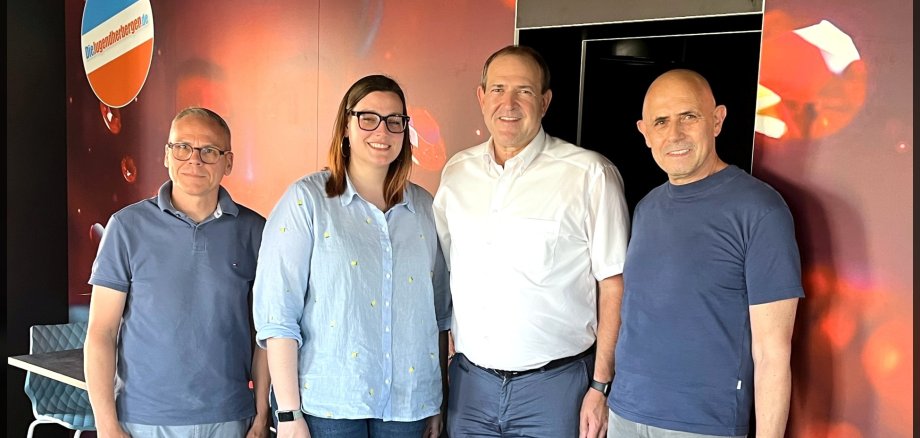 Auch im Restaurant ist das Edelsteinthema allgegenwärtig. Marco Böhm, Hannah Ihli, OB Frank Frühauf und Jacob Geditz (v. l.) freuen sich schon auf den Neustart der Edelsteinland-Jugendherberge. Das Foto zeigt die genannten Personen. Sie stehen nebeneinander im Restaurantbereich. Auf der Wand hinter Ihnen sind Edelsteine zu sehen.