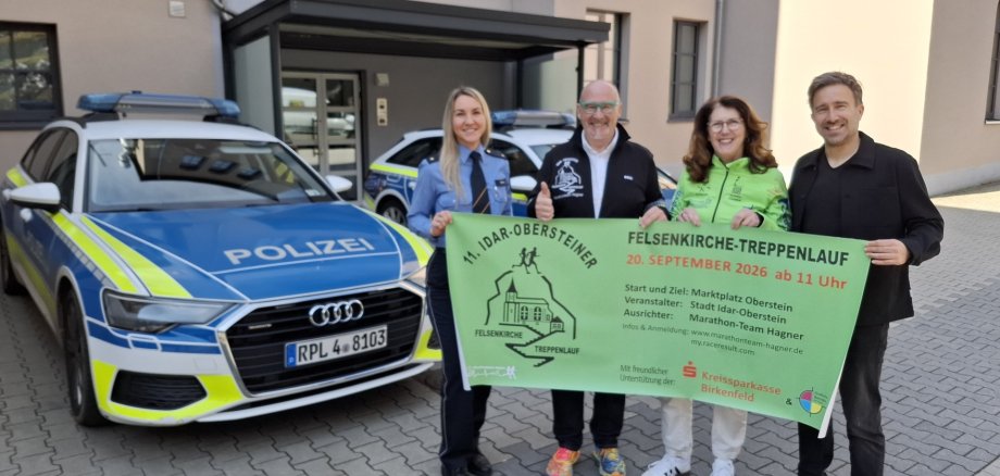 Die beiden Polizeichefs Kimberly Short (l.) und Timo Jung (r.) sagten Rainer und Ilonka Hagner Unterstützung für den Felsenkirche-Treppenlauf zu. Das Foto zeigt die genannten Personen im Hof der Polizeiinspektion. Sie stehen vor Streifenwagen und halten ein Banner des Treppenlaufs in die Kamera.